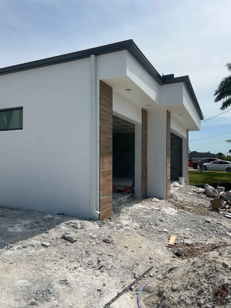 White stucco home addition with dark metal trim, wood doors, and integrated gutter system under construction